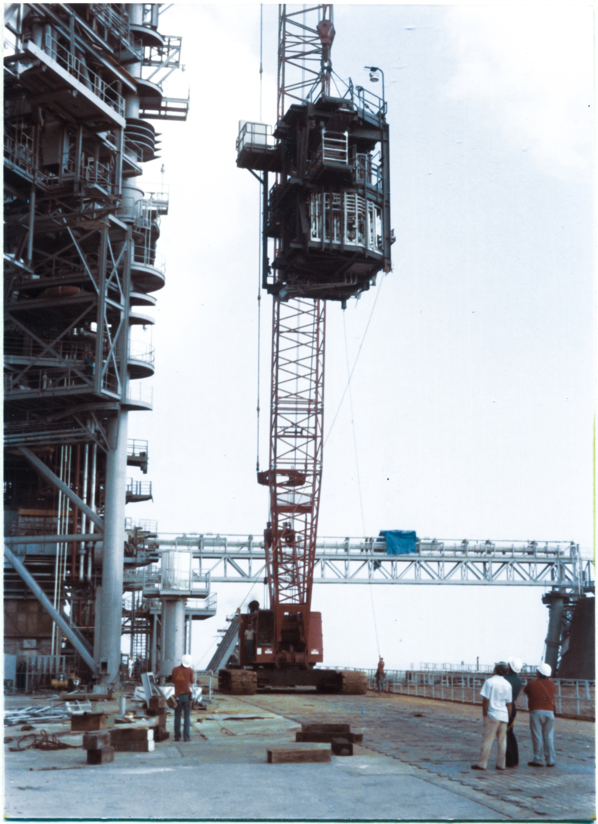 Image 091. Higher and higher into the air it goes. By this point in the OMBUU Lift at Space Shuttle Launch Complex 39-B, Kennedy Space Center, Florida, all of Ivey's Union Ironworkers involved with the lift and subsequent connection of the OMBUU to the face of the RSS had departed the Pad Deck, with the sole exception of the two who are working the tag lines. At this stage of things, I had moved closer to the lift with my camera, and also closer to the RSS, working to get a good angle on things, looking more directly upward at the OMBUU and also a bit east of due north. On the Pad Deck, Howard Baxter, recognizable by his red shirt and his build, stands with a small group of his TT&V people, watching the lift. In the distance, if you look close, you can see that Wade Ivey has climbed up on the crane, and is standing right next to the crane operator, eyeing things closely, staying right on top of things as they unfold. Photo by James MacLaren.